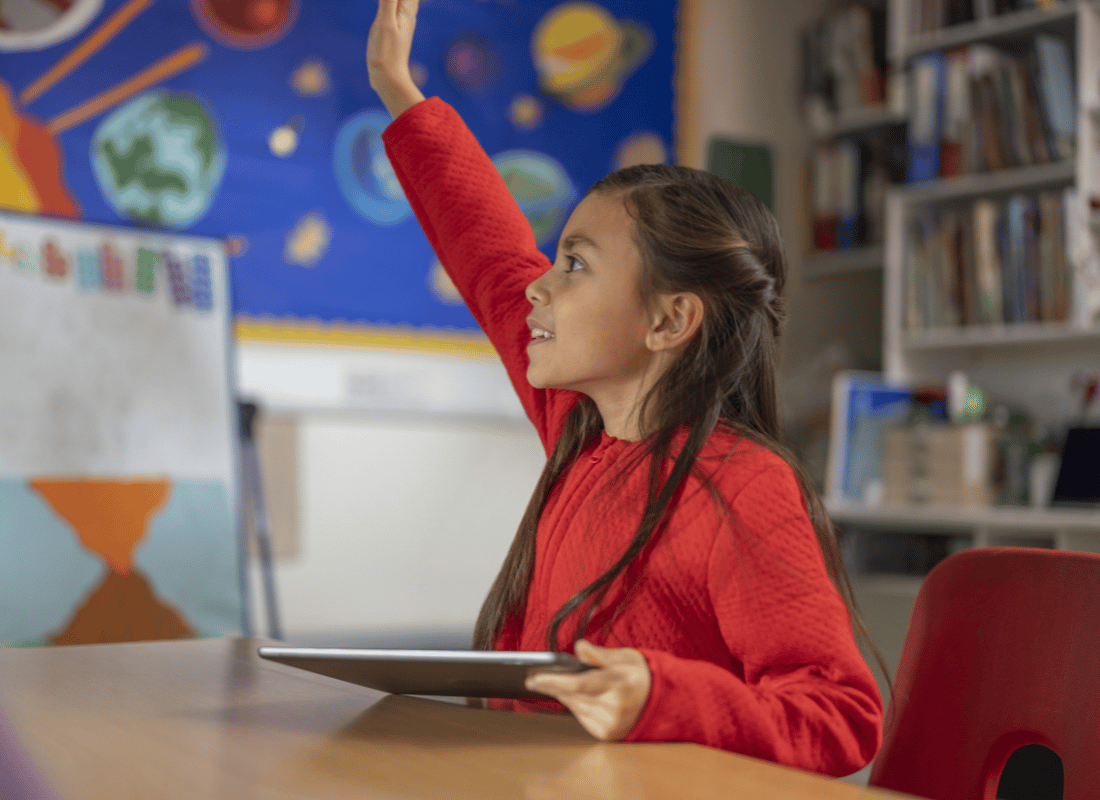 Keen primary school student holding an iPad, raises hand in class