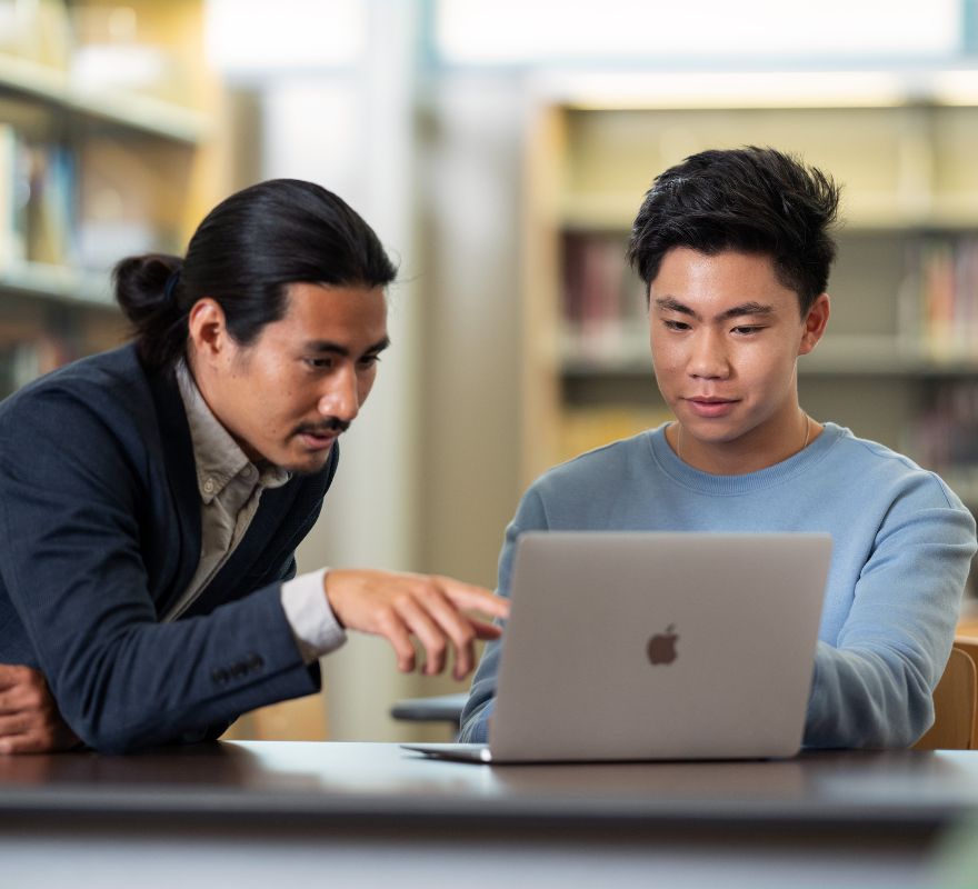 Two people working together in a library whilst using a Mac.